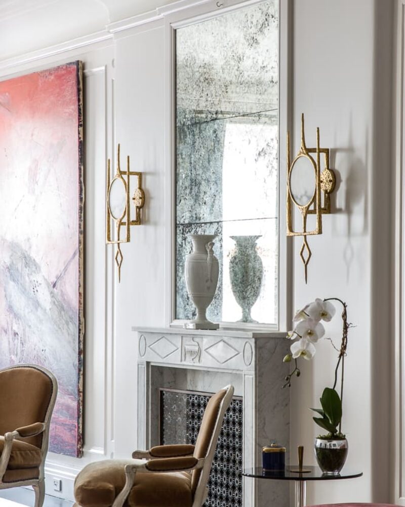 A refined living space featuring a marble fireplace, antique mirror, gold sconces, plush velvet chairs, and a striking abstract painting.