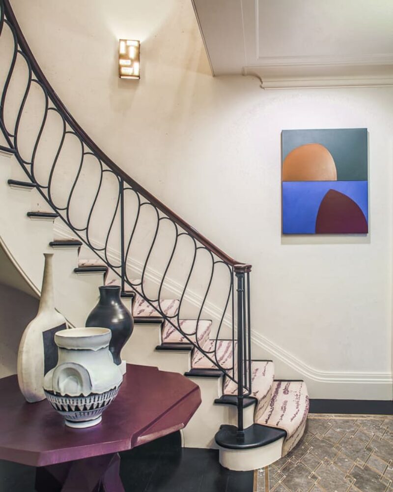 A striking staircase with an elegant curved railing, geometric side table with sculptural vases, and a bold modern painting on the wall.
