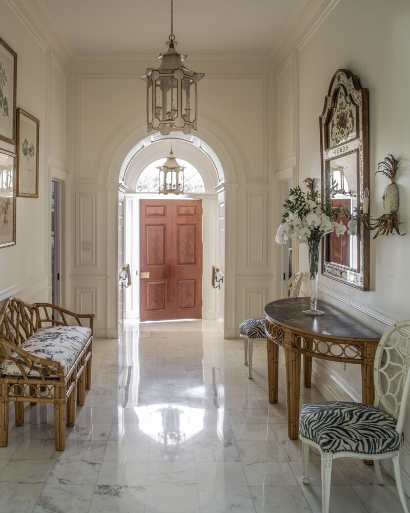 Elegant hallway with marble floors, arched wooden door, ornate mirror, and stylish furniture under a vintage chandelier.
