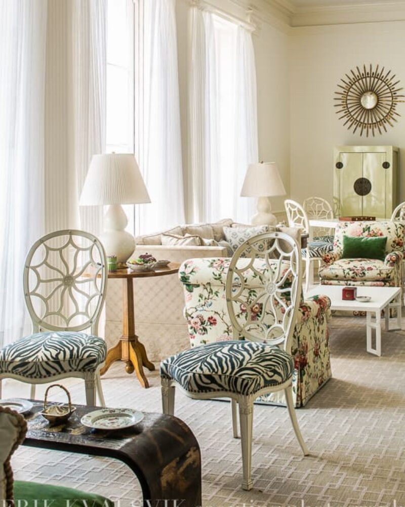 Elegant living room with floral sofas, zebra-patterned chairs, sunburst mirror, and soft lighting from large windows.