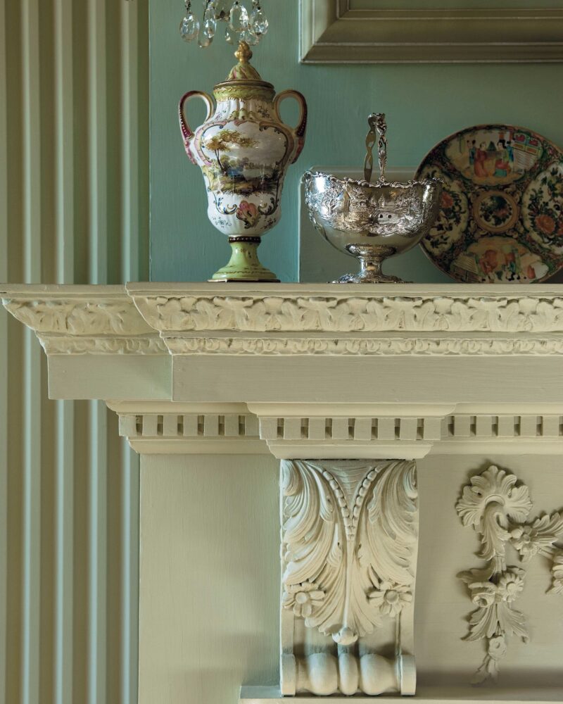 Elegant mantel with ornate vase, silver bowl, and decorative plate, framed by intricate woodwork and chandelier.