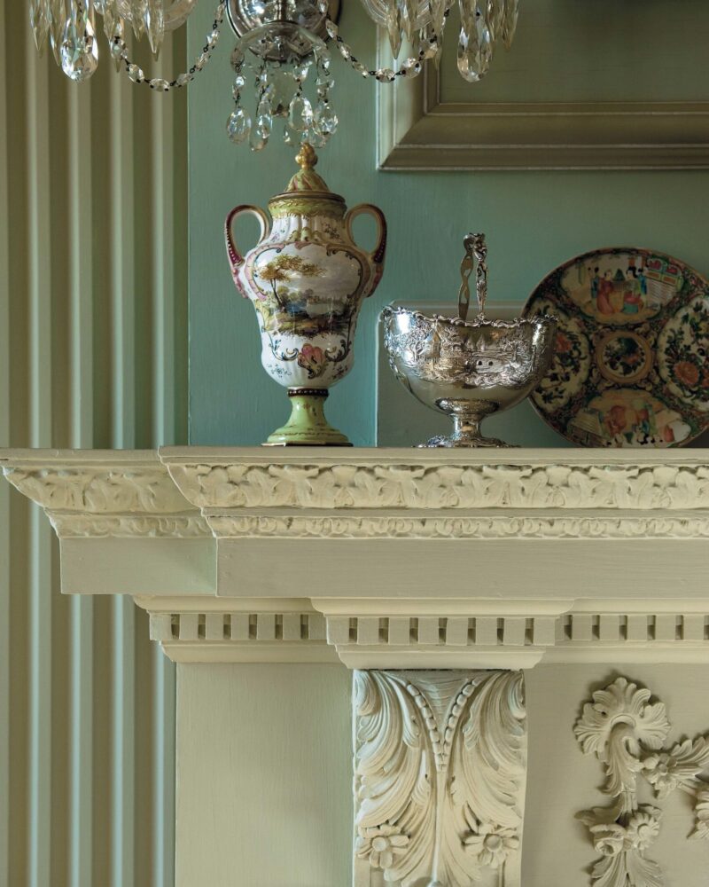 Elegant mantel with ornate vase, silver bowl, and decorative plate, framed by intricate woodwork and chandelier.