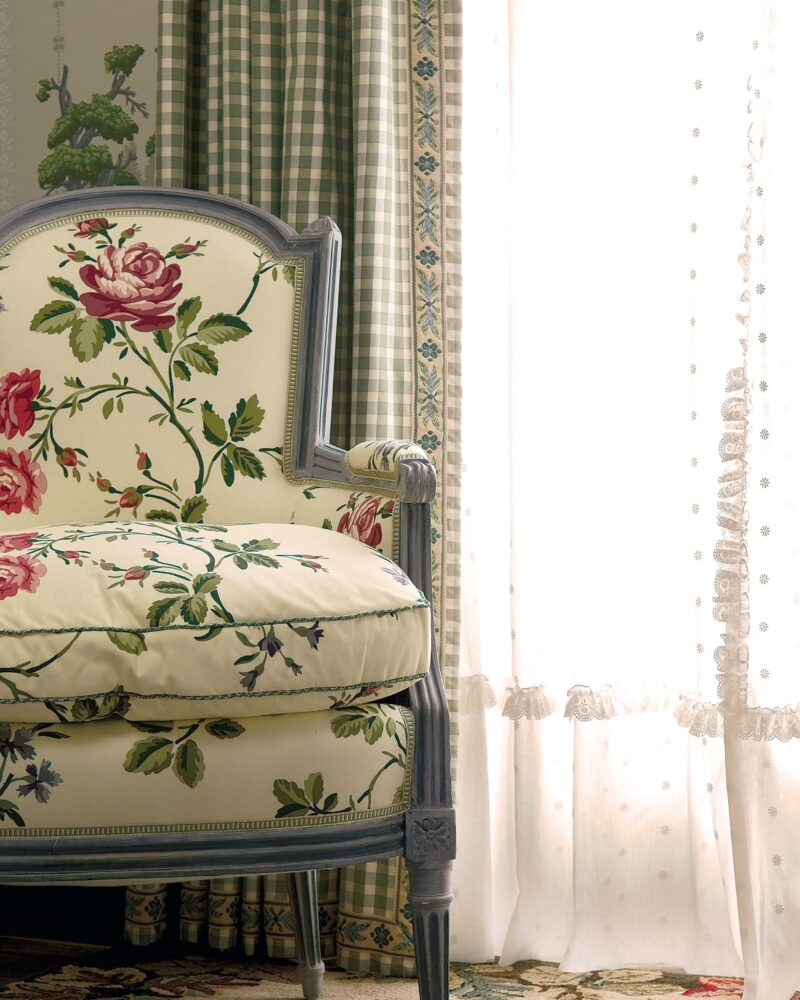 Floral armchair with green checkered curtains and sheer lace by a sunlit window, set on a patterned rug.