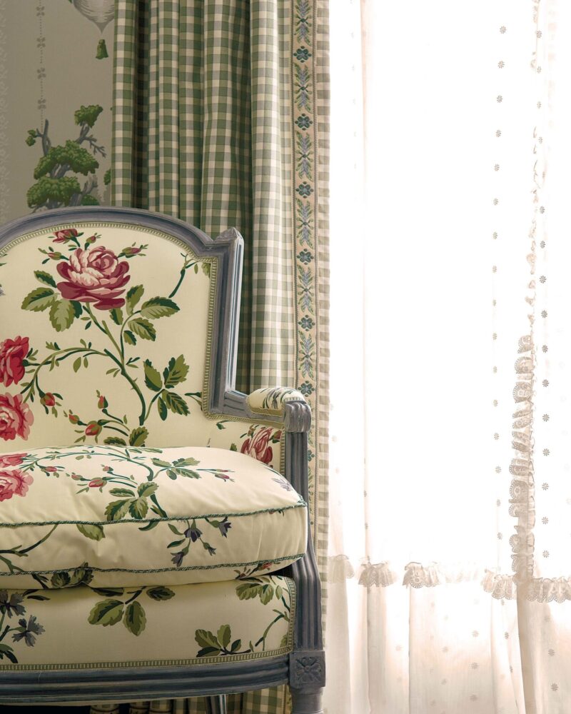 Floral armchair with green checkered curtains and sheer lace by a sunlit window, set on a patterned rug.