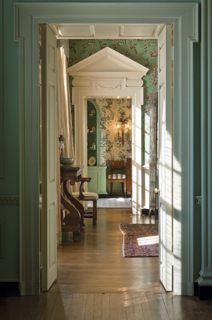 Elegant hallway with floral wallpaper, wooden floors, and classic furniture, illuminated by soft natural light.