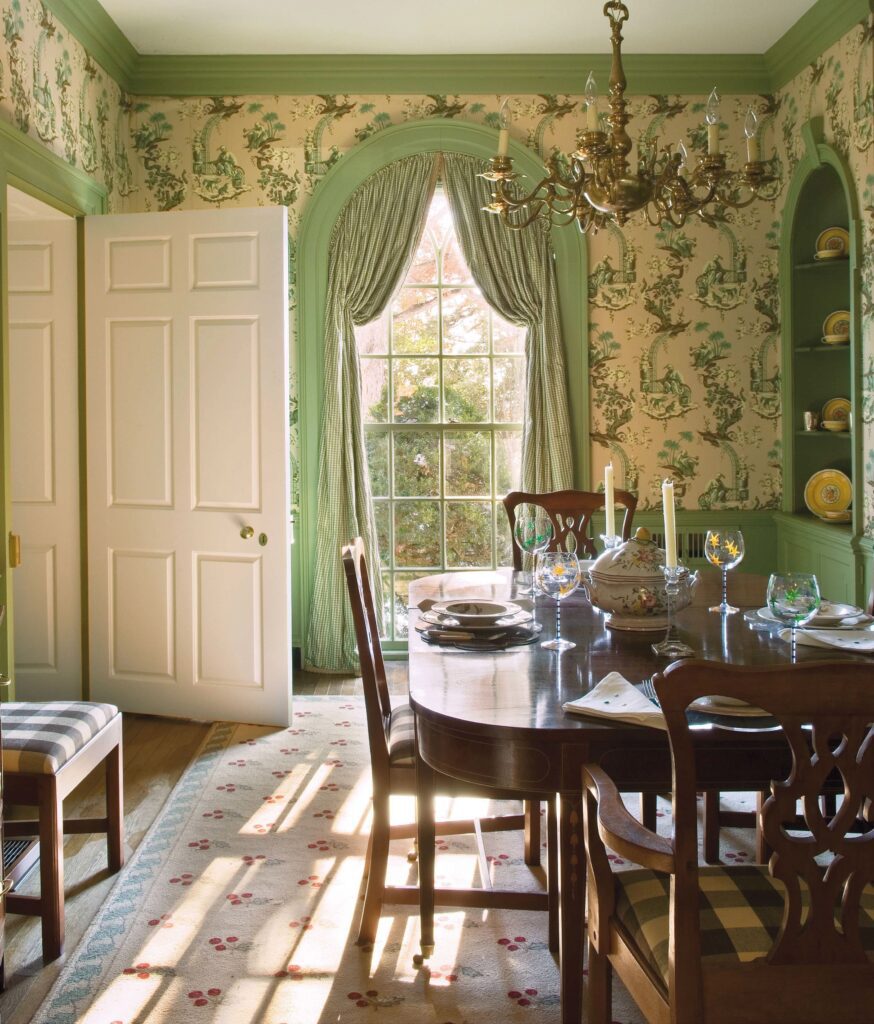 Elegant dining room with green accents, floral wallpaper, arched window, and chandelier. Sunlight streams onto the table setting.
