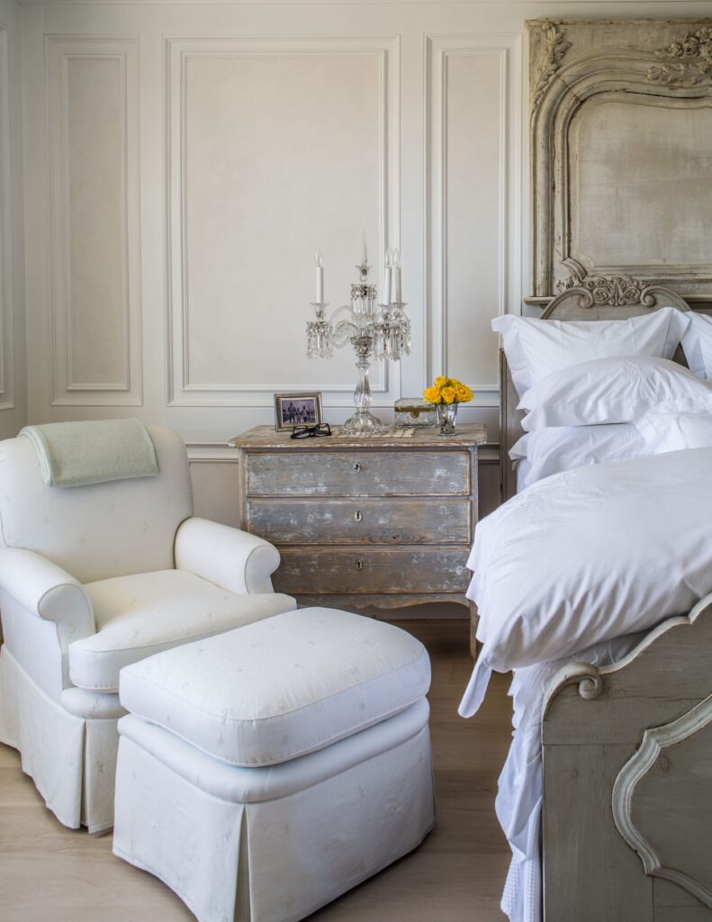 Elegant bedroom with vintage armchair, ornate bed, distressed dresser, and crystal candelabra.