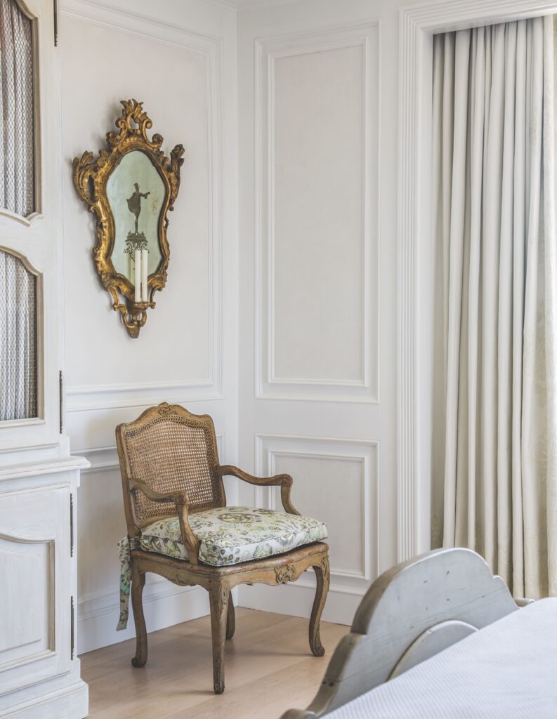 Elegant room with ornate mirror, vintage chair with floral cushion, and white paneled walls, exuding classic charm.