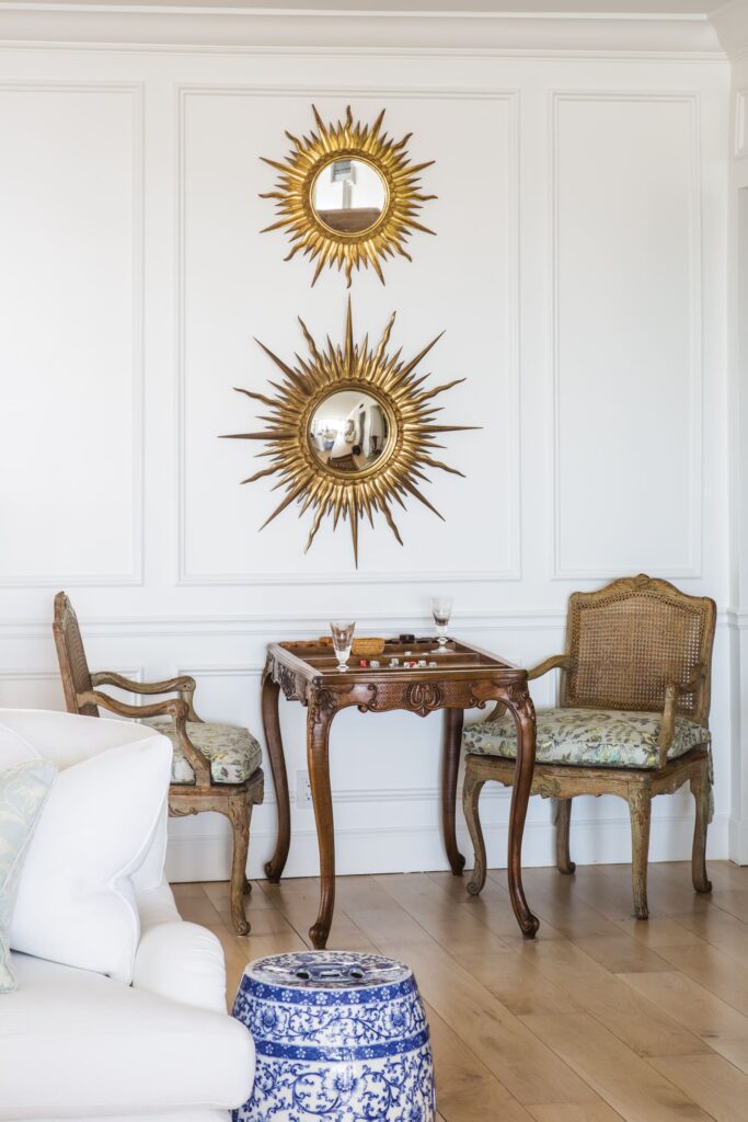 Elegant room with sunburst mirrors, vintage chairs, a wooden table, and a blue ceramic stool on wooden flooring.