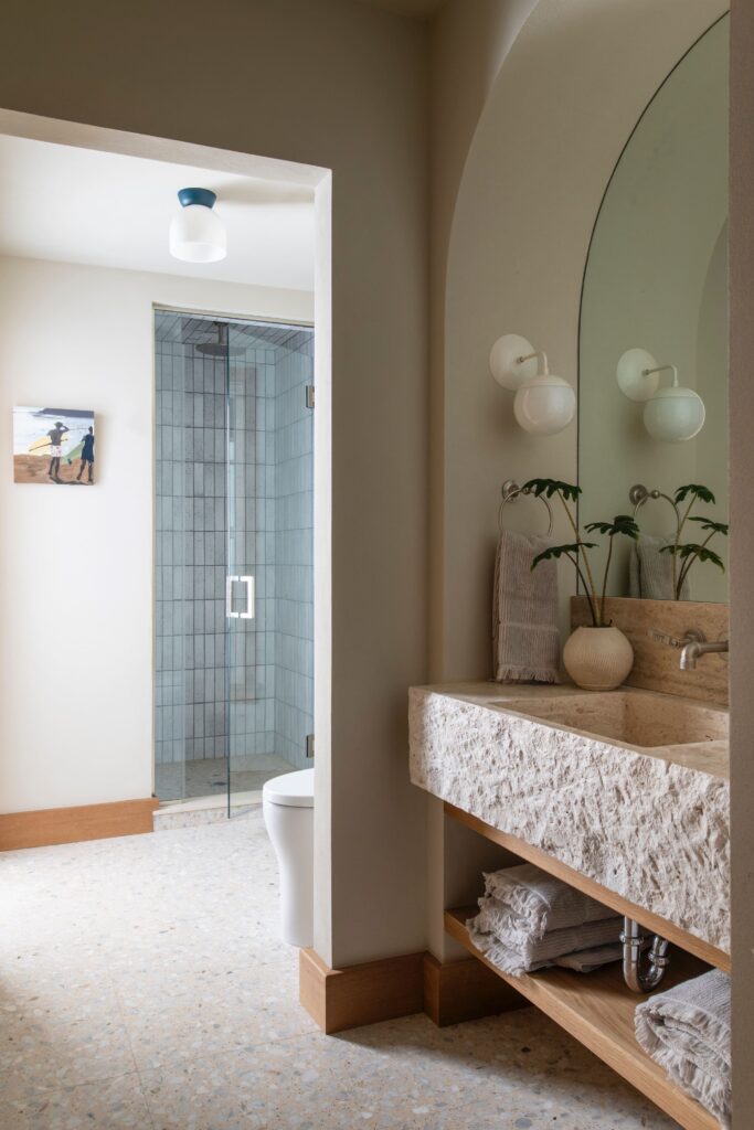 Modern bathroom with stone sink, large mirror, glass shower, and plant decor. Light walls and terrazzo flooring enhance elegance.