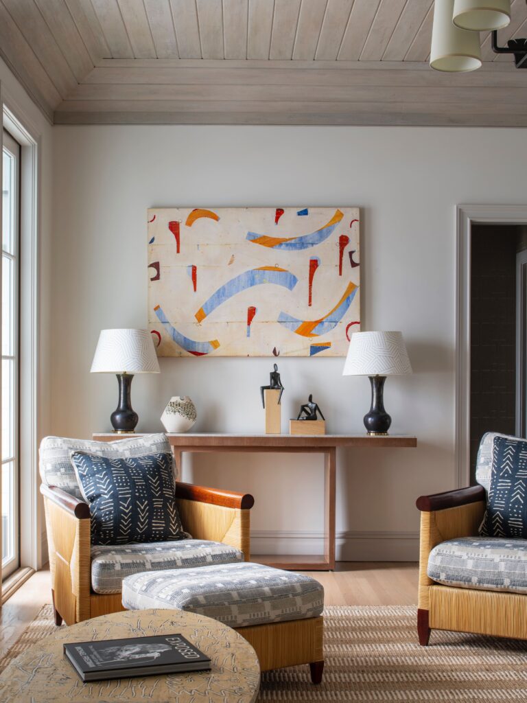 Modern living room with abstract art, patterned chairs, and a wooden table with sculptures. Natural light from large window.