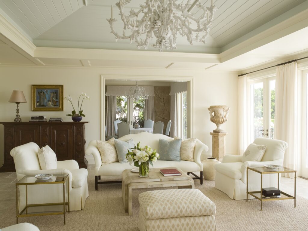 A refined living room with a soft neutral palette, sculptural white chandelier, plush seating, antique furnishings, and natural light streaming through French doors.