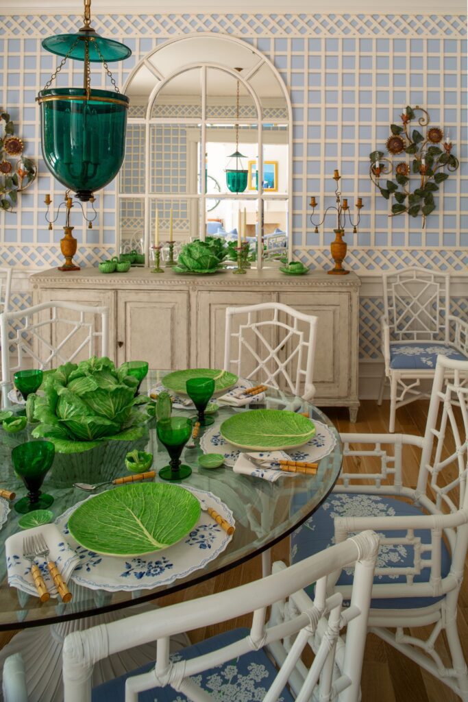 Elegant dining room with green glassware, leaf-patterned plates, and white chairs against a blue lattice wall.