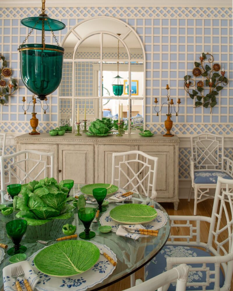 Elegant dining room with green glassware, leaf-patterned plates, and white chairs against a blue lattice wall.