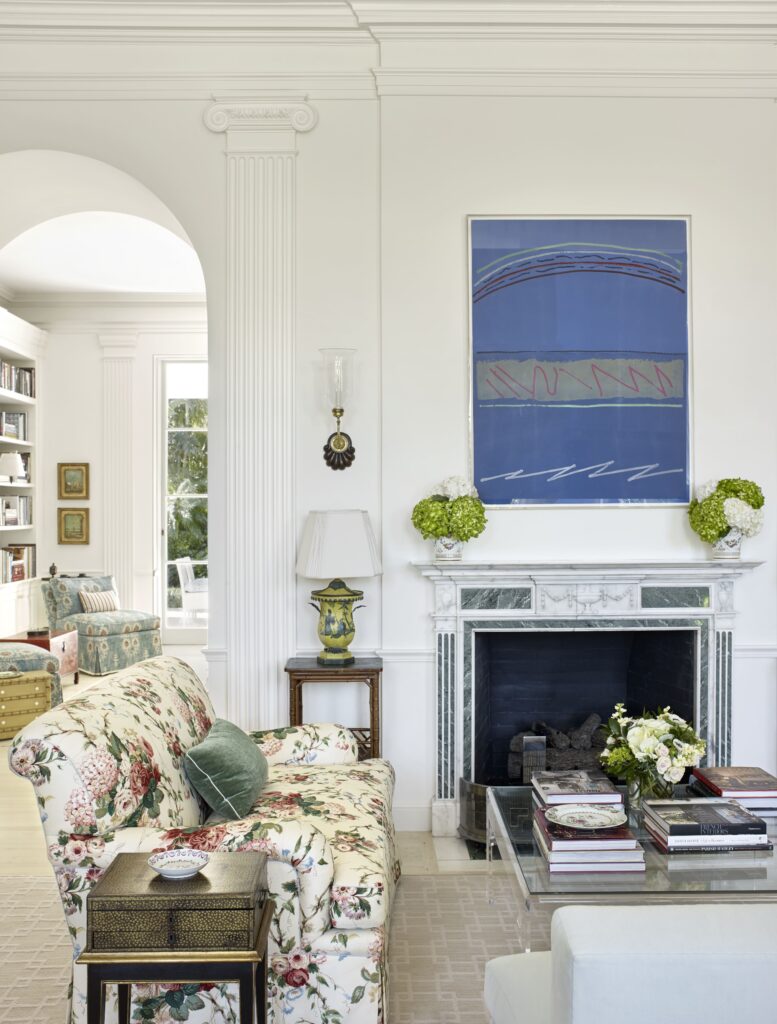 Elegant living room with floral sofa, marble fireplace, abstract art, and books on a glass coffee table.