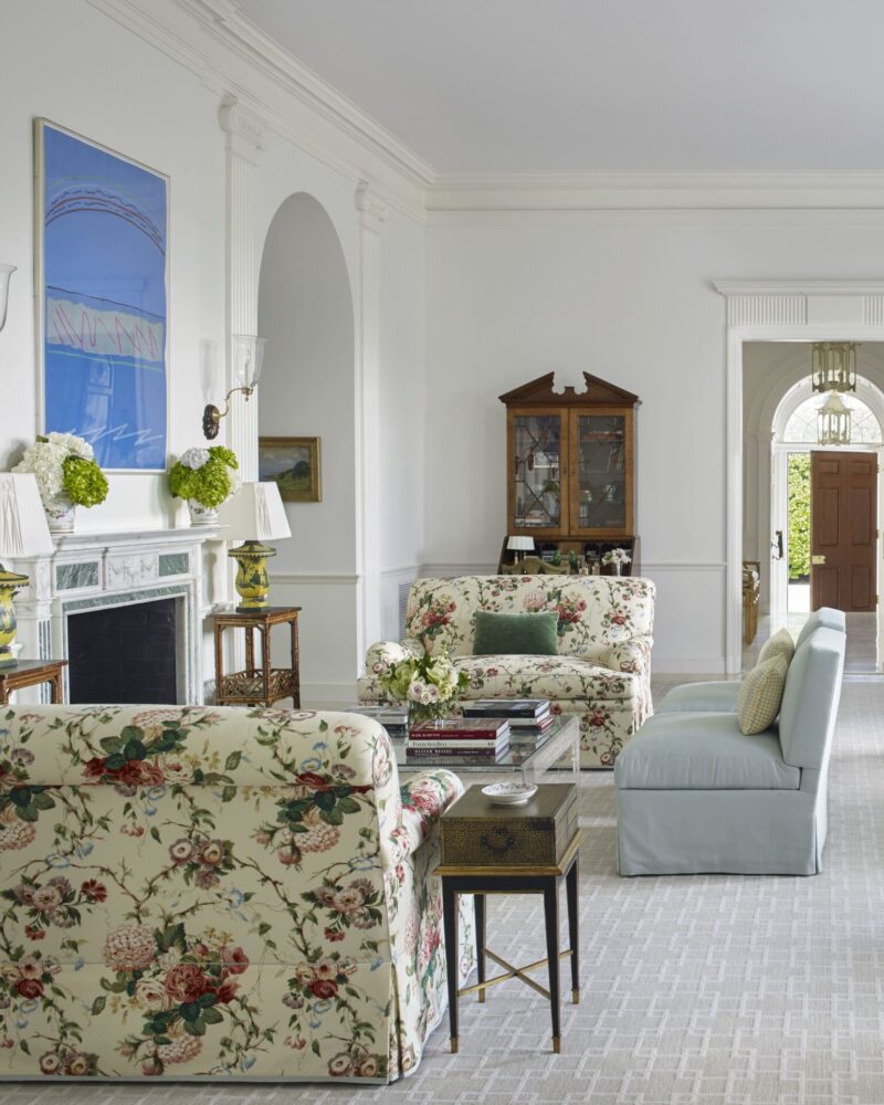 Elegant living room with floral furniture, light blue sofa, and a classic fireplace. Bright, airy space with large windows.