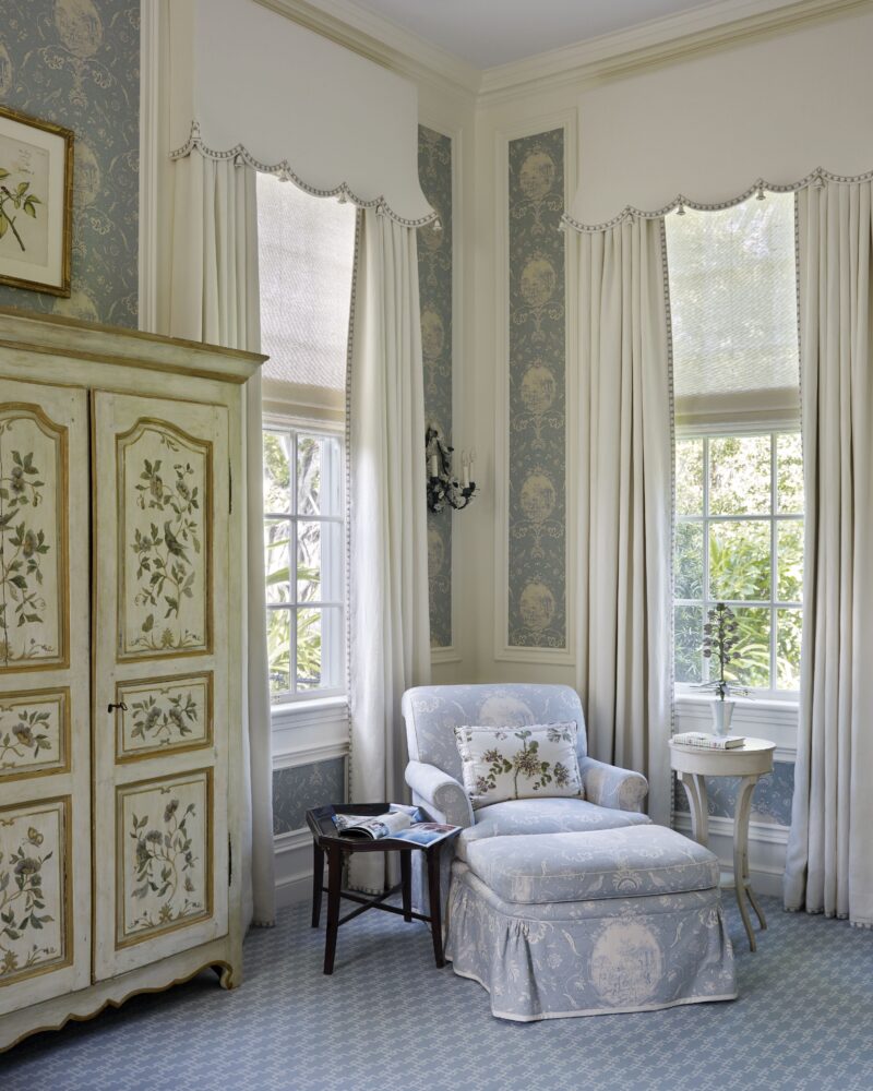 Elegant sitting area with floral armchair, matching ottoman, vintage cabinet, and decorative wallpaper near large windows.
