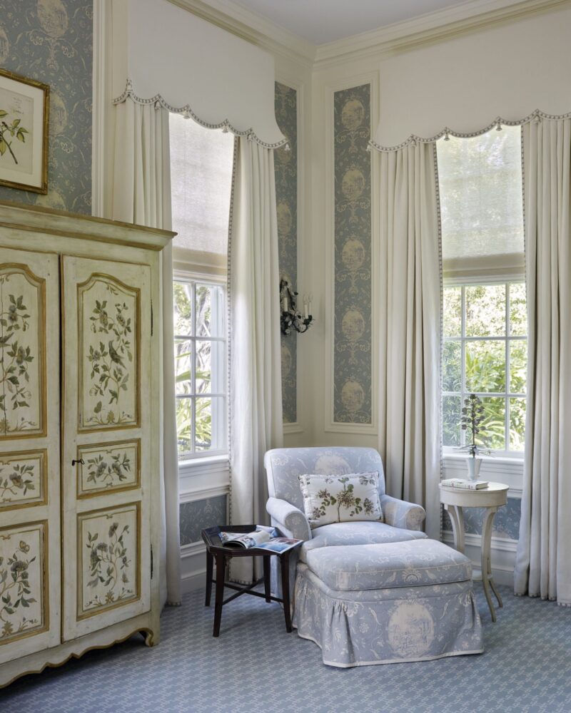 Elegant sitting area with floral armchair, matching ottoman, vintage cabinet, and decorative wallpaper near large windows.