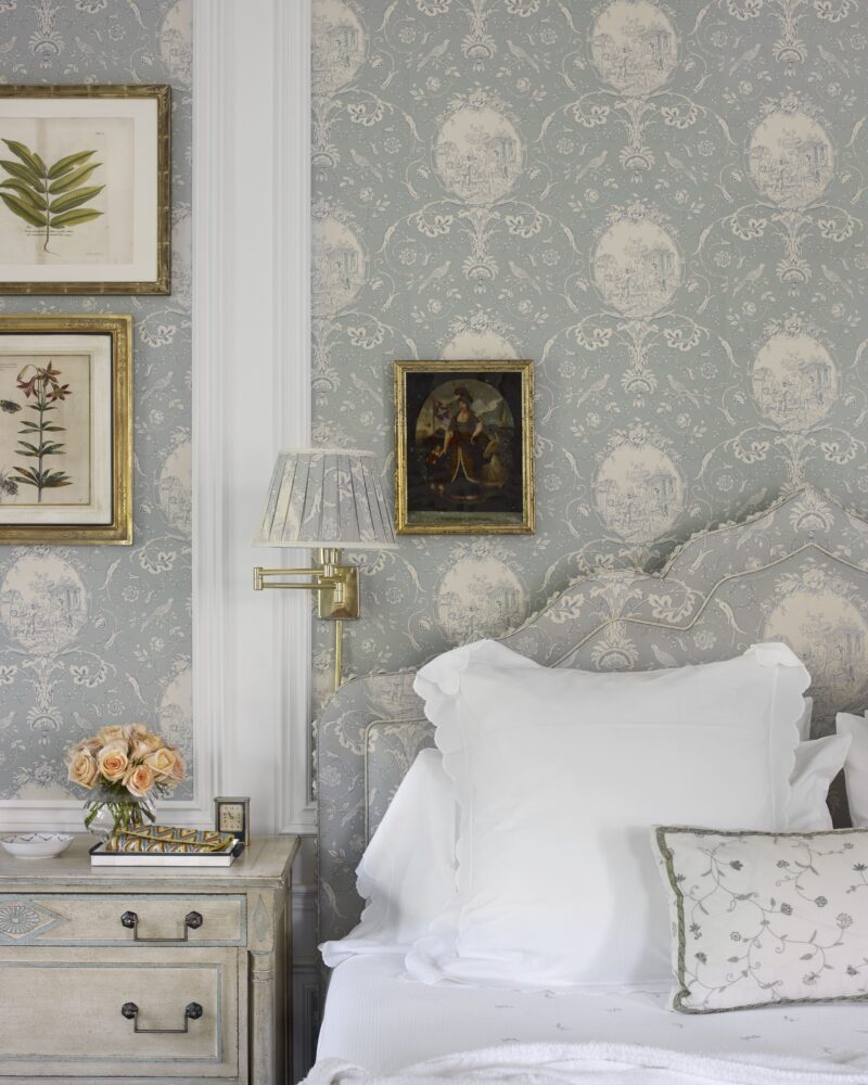 Elegant bedroom with floral wallpaper, white bedding, vintage nightstand, framed botanical prints, and a vase of roses.