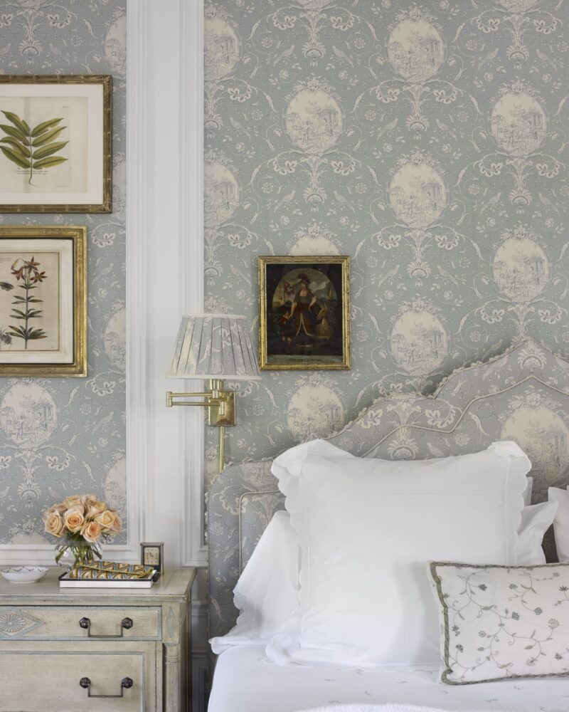 Elegant bedroom with floral wallpaper, white bedding, vintage nightstand, framed botanical prints, and a vase of roses.