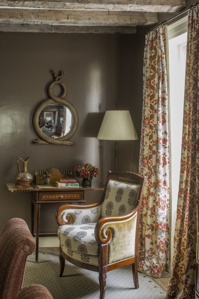 Vintage armchair and ornate mirror in a cozy room with floral curtains and a wooden table.