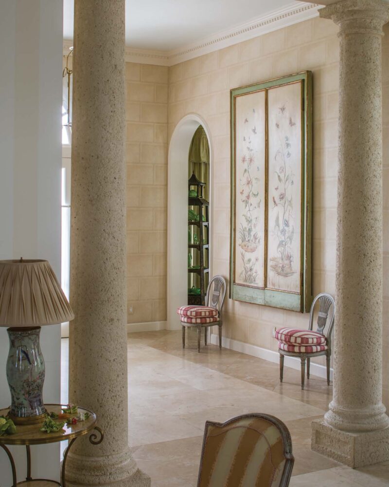 Elegant interior with stone columns, striped chairs, and floral artwork on a beige wall. Marble flooring adds sophistication.