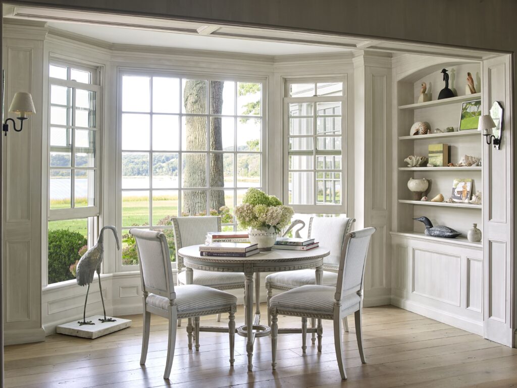 Elegant sunlit room with a round table, white chairs, large windows, and decorative shelves overlooking a scenic landscape.
