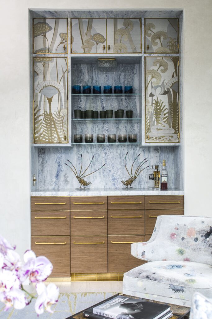 Elegant home bar with decorative cabinetry, glass shelves holding cups, wooden drawers, and a patterned chair.