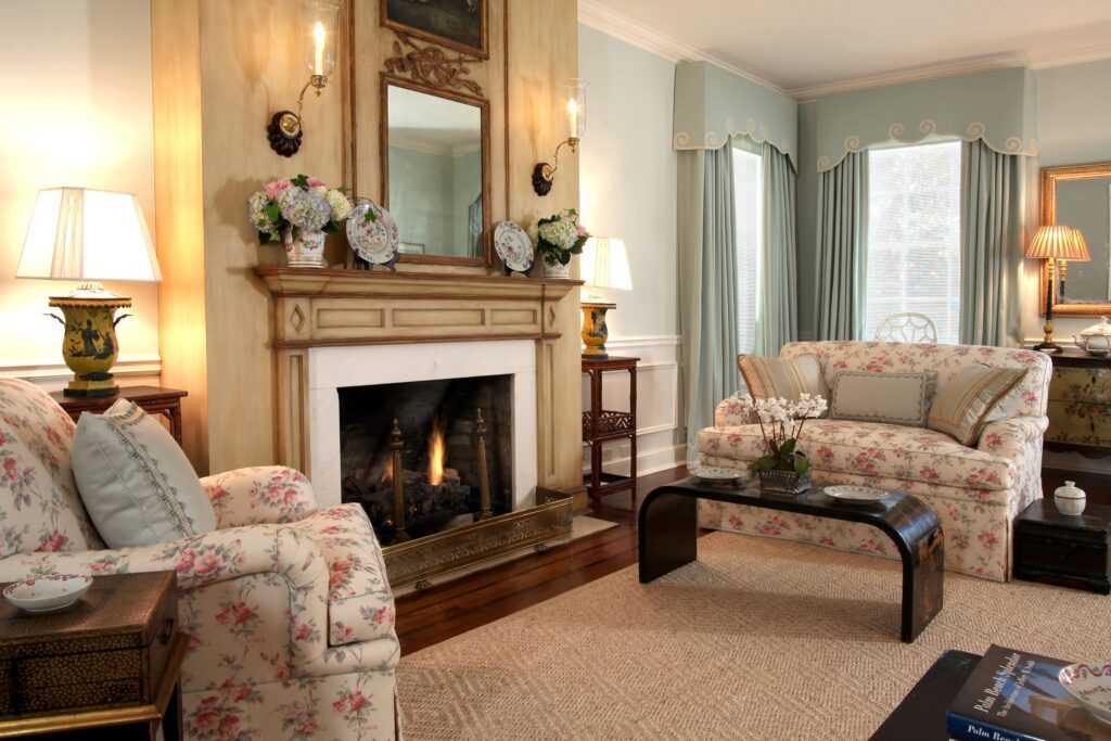 An elegant living room with a classic design, featuring a warm fireplace framed by an ornate wooden mantel with a mirror and decorative plates. Floral upholstered sofas with decorative pillows complement the soft blue walls and drapery with intricate trim. A woven jute rug anchors the space, while antique-style lamps and a dark wood coffee table add sophistication. The room is illuminated by natural light streaming through large windows.