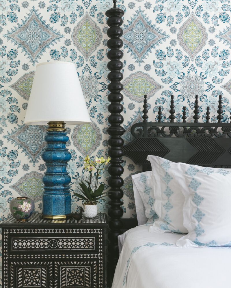 Ornate bedroom with patterned wallpaper, intricate nightstand, and blue lamp beside a bed with decorative pillows.