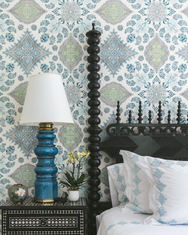 Ornate bedroom with patterned wallpaper, intricate nightstand, and blue lamp beside a bed with decorative pillows.