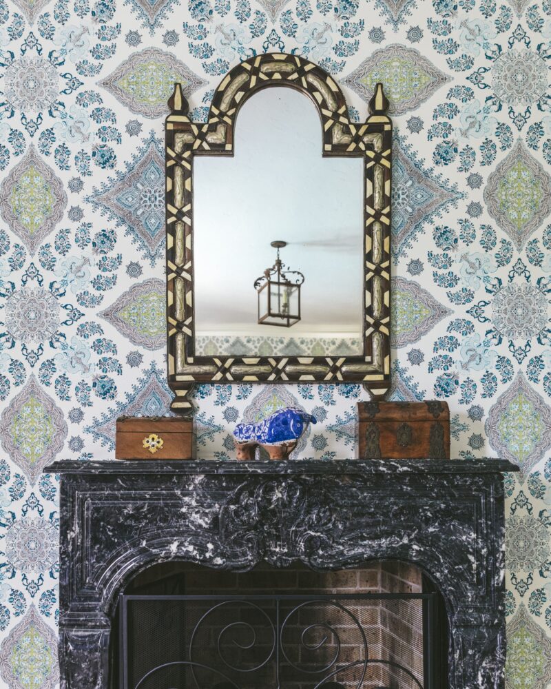 Ornate mirror above a black marble fireplace with intricate patterned wallpaper and decorative wooden boxes.