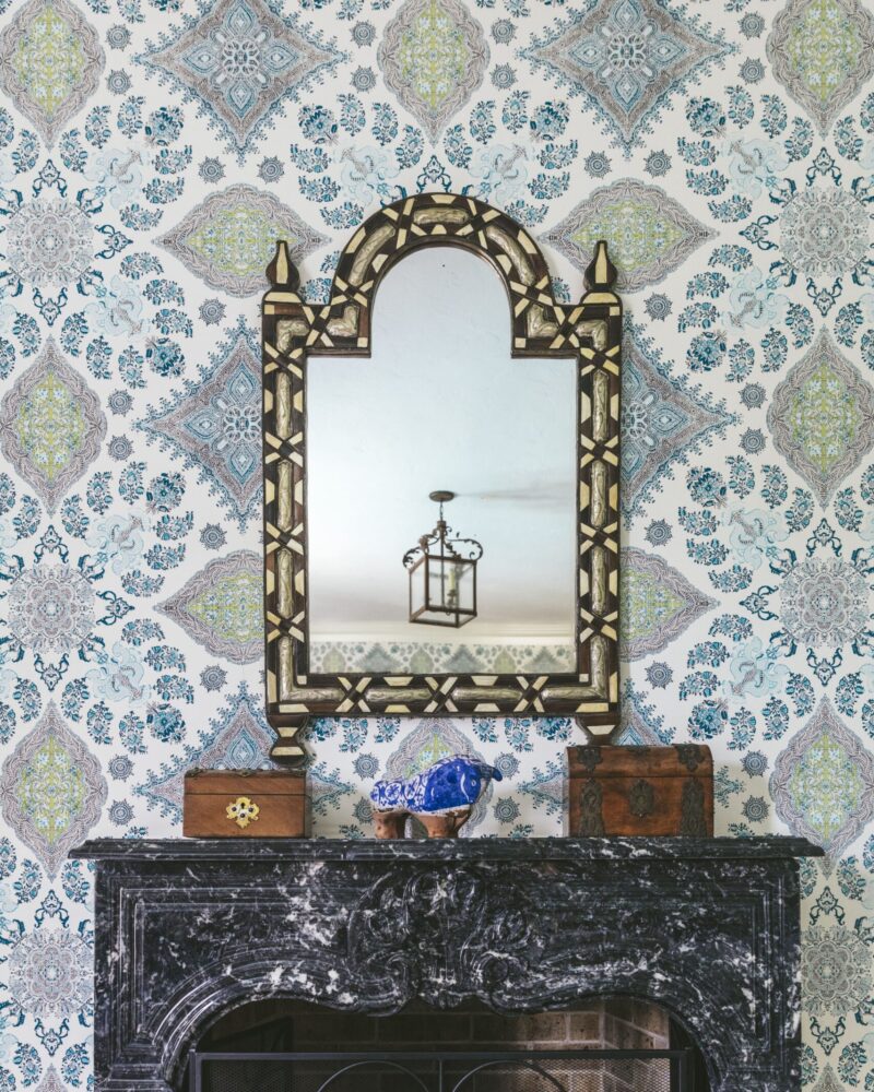 Ornate mirror above a black marble fireplace with intricate patterned wallpaper and decorative wooden boxes.
