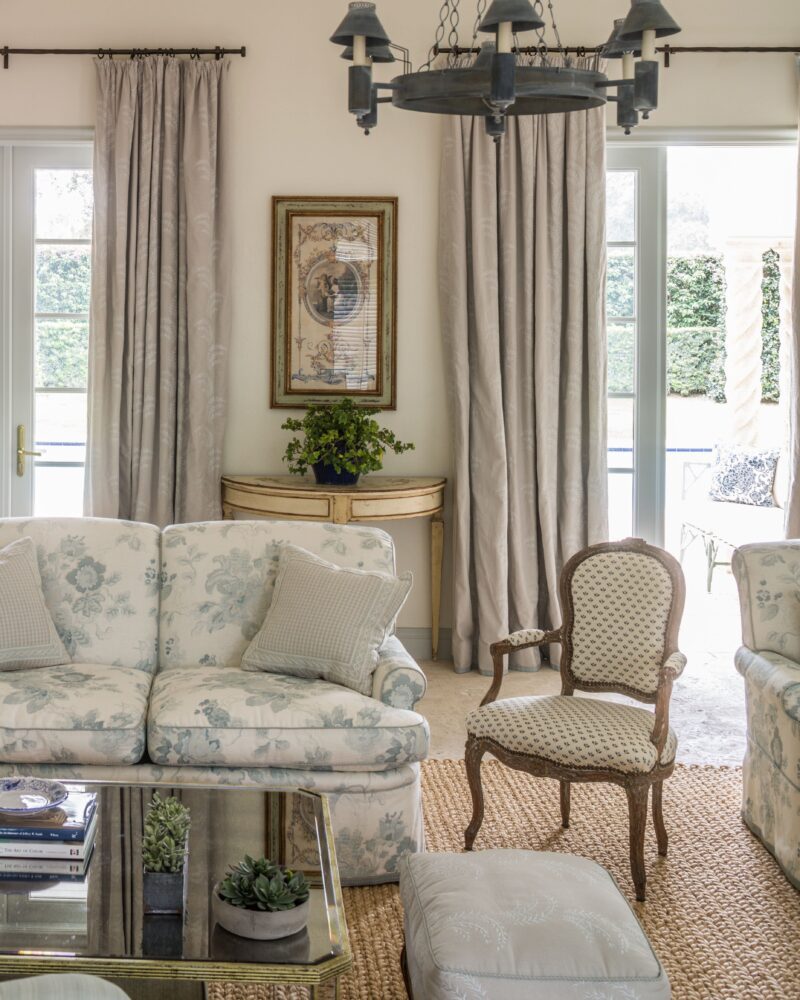 Elegant living room with floral sofa, vintage armchair, glass coffee table, and chandelier. Neutral tones and classic decor.