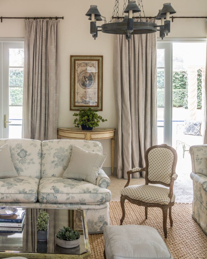 Elegant living room with floral sofa, vintage armchair, glass coffee table, and chandelier. Neutral tones and classic decor.
