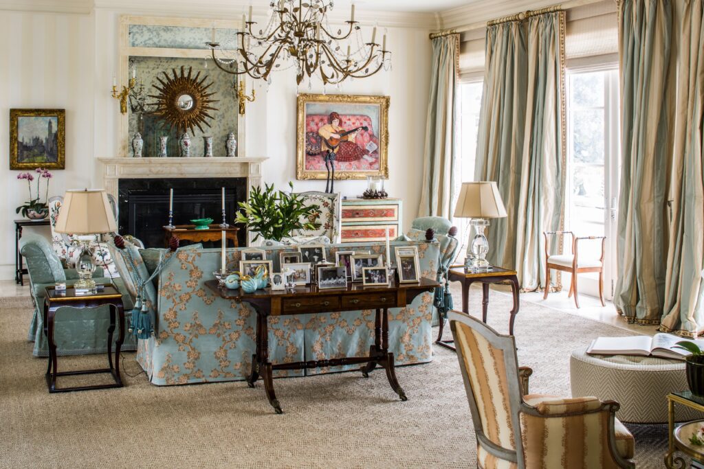 Elegant living room with ornate chandelier, floral sofas, framed photos, and large windows with flowing curtains.
