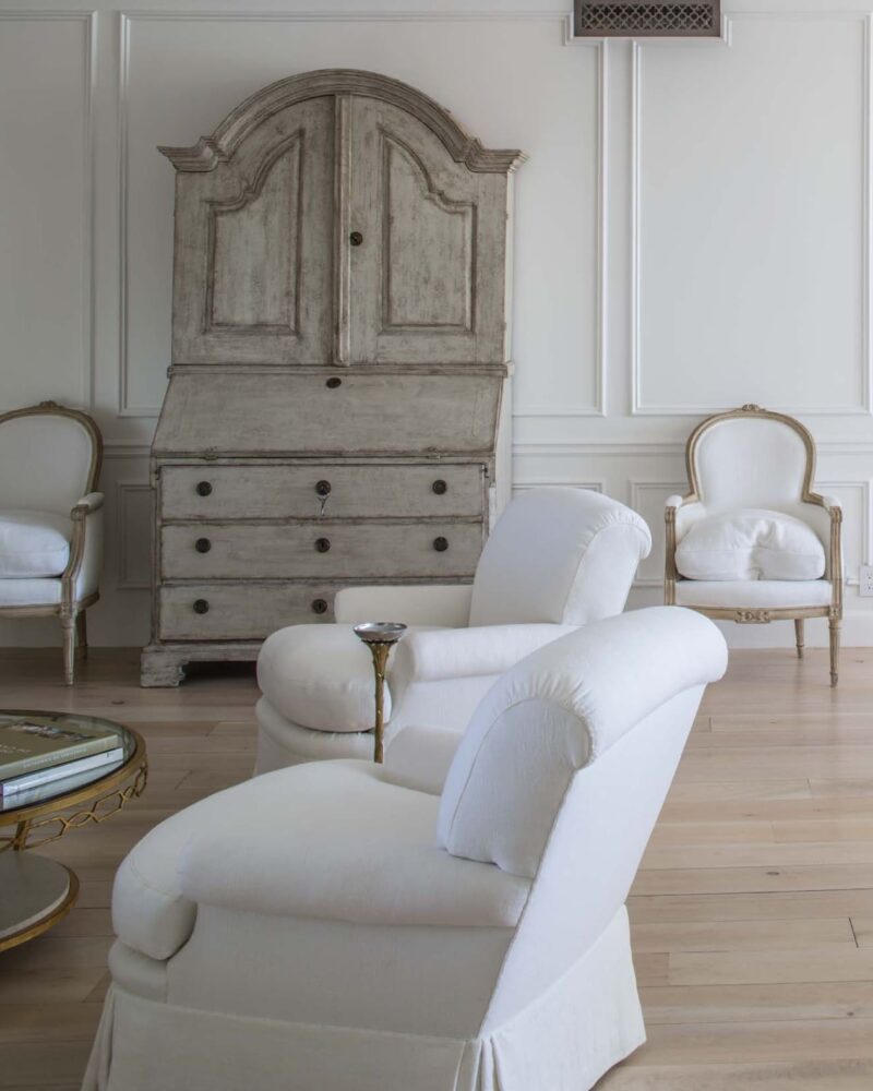 Elegant living room with white armchairs, a vintage wooden cabinet, and a glass coffee table on light wood flooring.