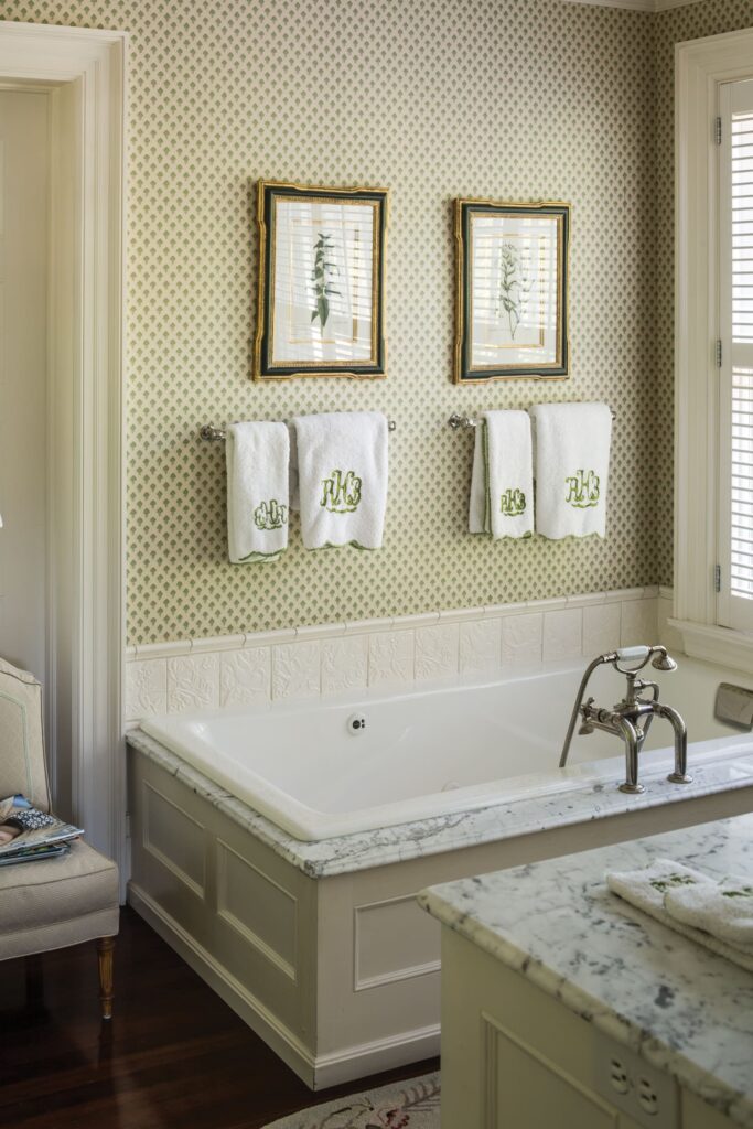 Elegant bathroom with patterned wallpaper, framed botanical art, and monogrammed towels above a marble-topped bathtub.