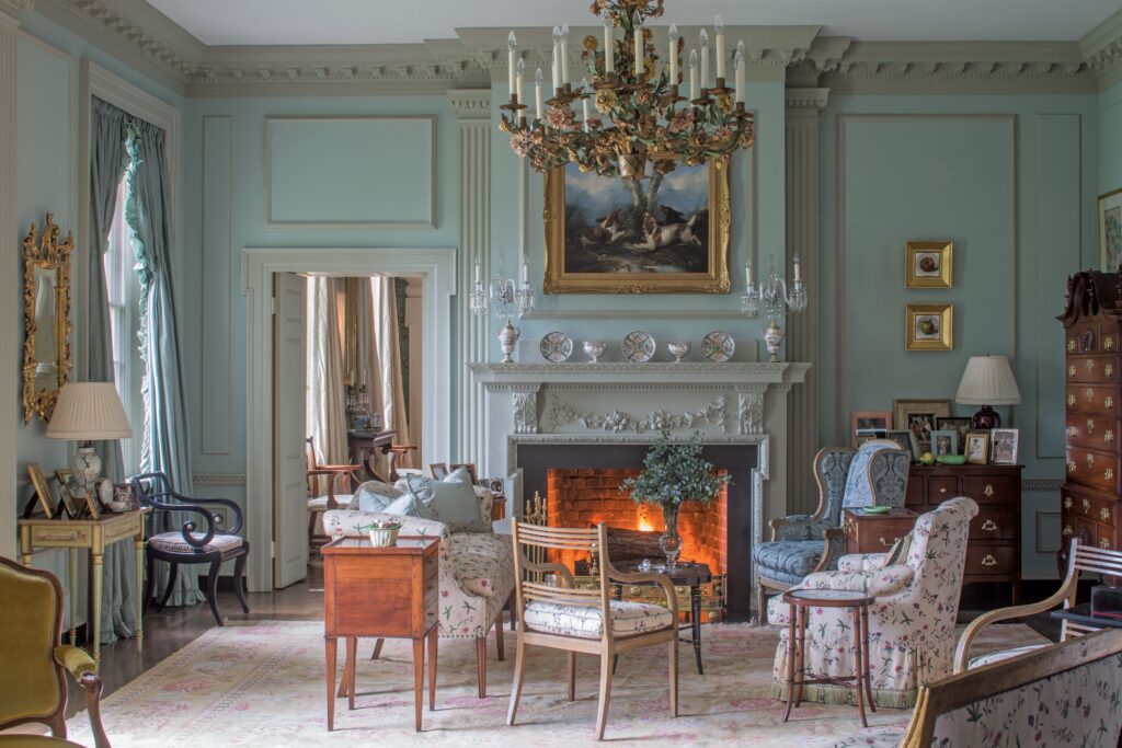 A classic, elegant living room with a lit fireplace, antique furniture, floral upholstery, and a grand chandelier in a soft blue-green setting.