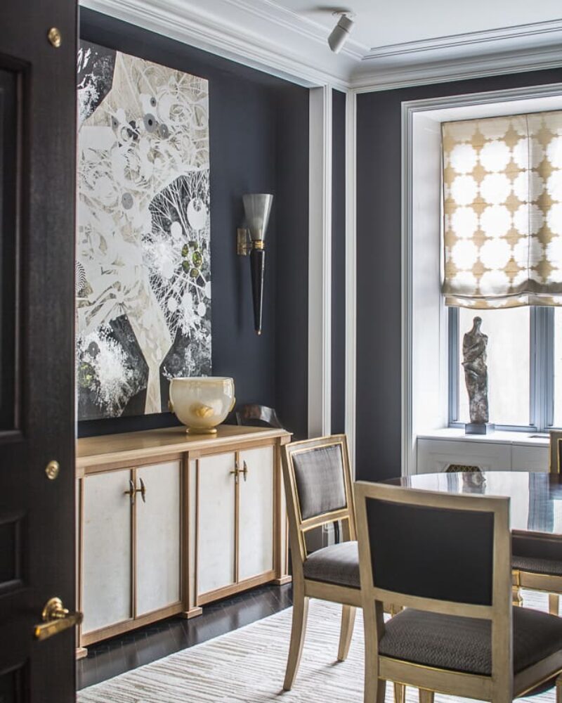A sophisticated dining room with dark walls, gold-accented furniture, abstract artwork, and a sculptural centerpiece by the window.