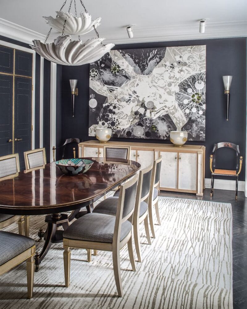 Elegant dining room with dark walls, abstract artwork, chandelier, wooden table, upholstered chairs, and patterned rug.