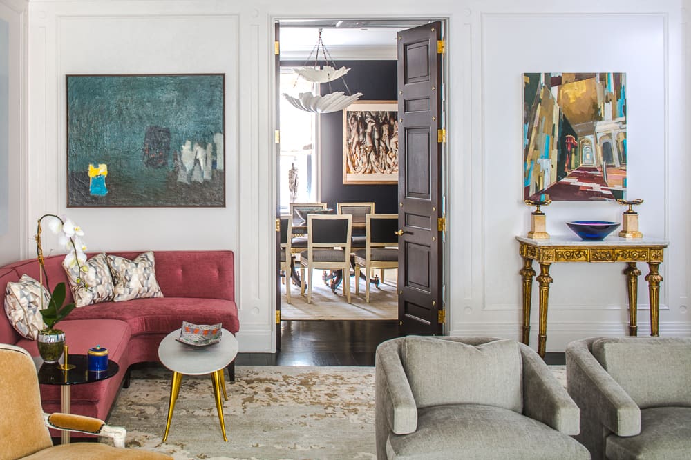 Elegant living room with red sofa, abstract paintings, and view into a dining area through dark wood doors.