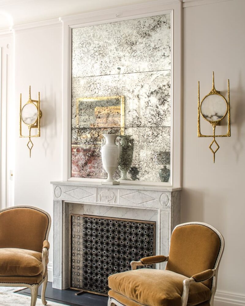 A sophisticated living space with a marble fireplace, antique mirror, gold sconces, and plush velvet chairs in a refined neutral palette.