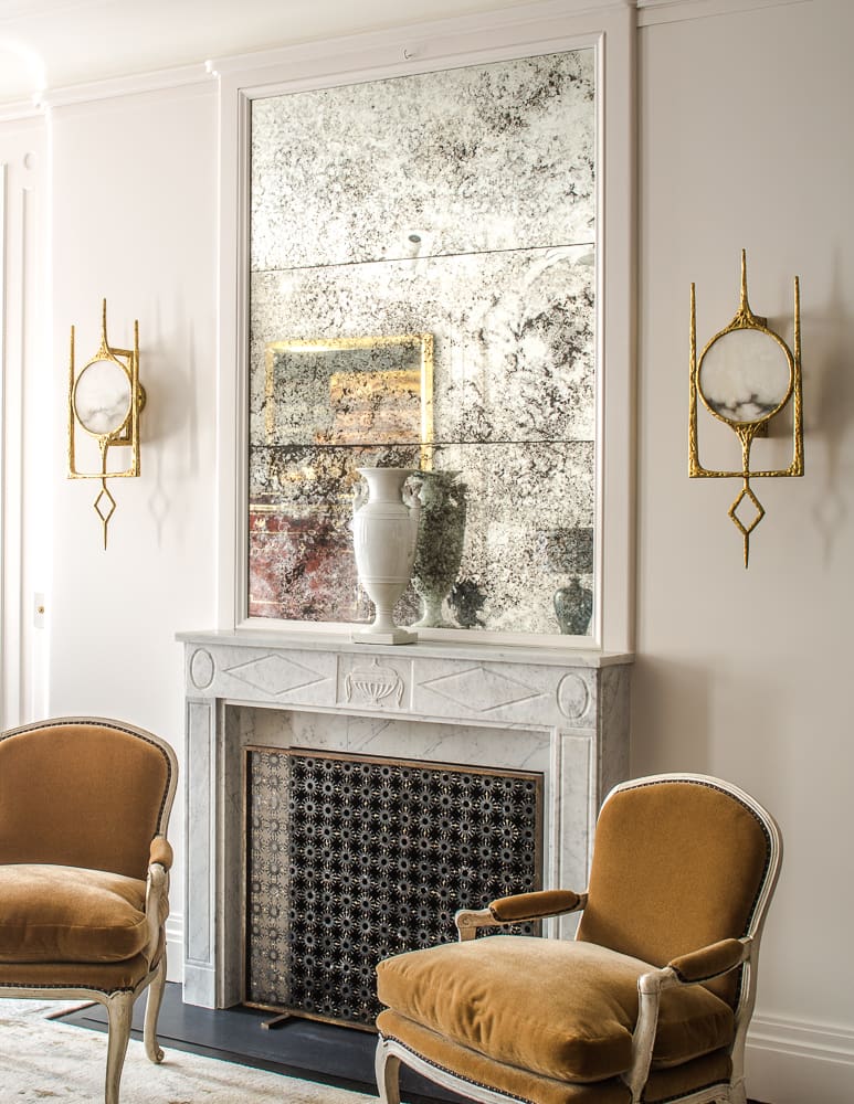 A sophisticated living space with a marble fireplace, antique mirror, gold sconces, and plush velvet chairs in a refined neutral palette.
