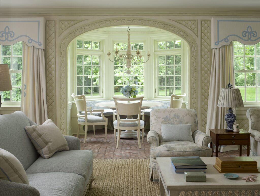 Elegant living room with floral armchairs, a cozy sofa, and a sunlit bay window dining area featuring a chandelier.
