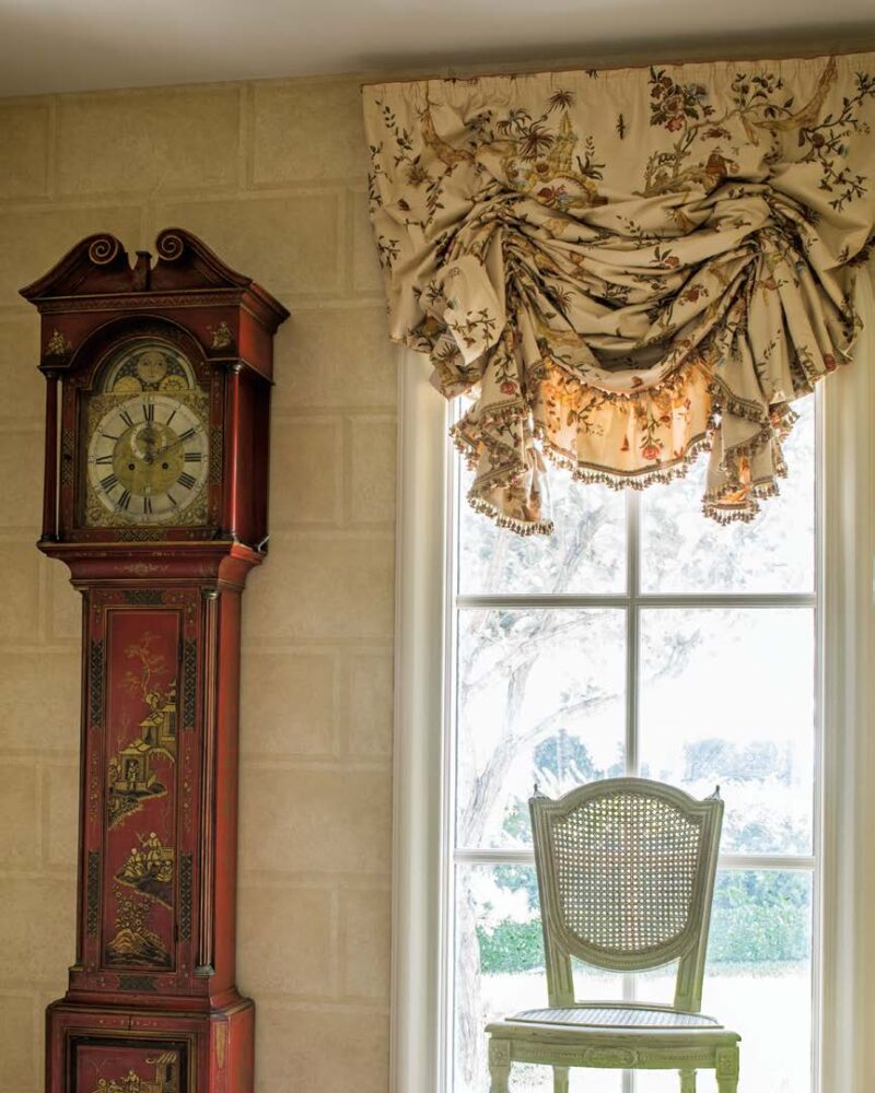 Elegant room with vintage grandfather clock, ornate drapery, and a classic chair by a sunlit window.