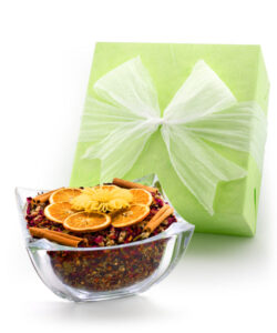 A glass bowl filled with potpourri, topped with orange slices and cinnamon sticks, next to a green gift box.