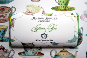 Milk chocolate bar labeled "Green Tea" with decorative packaging, set against a background of colorful teacups.