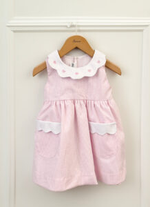 A pink striped dress with a white scalloped collar and pockets hangs on a wooden hanger.