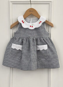 A black and white gingham dress with a white collar and cherry embroidery, hanging on a wooden hanger.